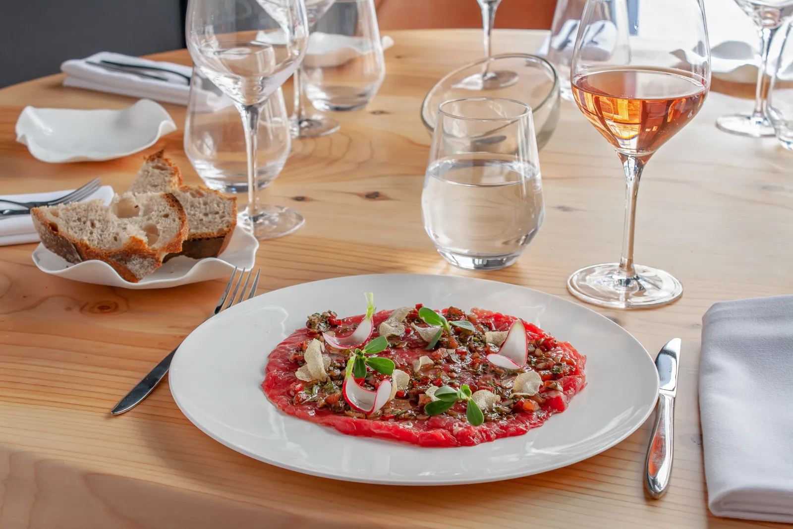 Plate of beef carpaccio topped with pistachios, radish slices and microgreens, served on a wooden table with wine glasses, toasted bread and cutlery at Le Loup restaurant in Bordeaux