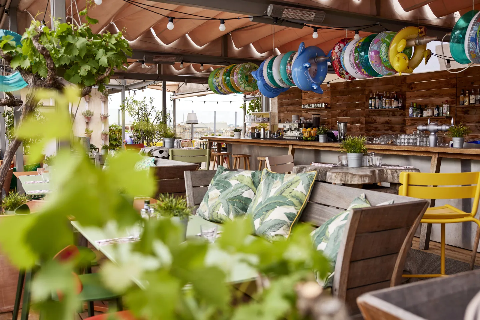 Interior of Mama Shelter Bordeaux restaurant featuring a covered terrace with hanging plants, colorful decorative plates on the ceiling, wooden furnishings, and views of Bordeaux cityscape