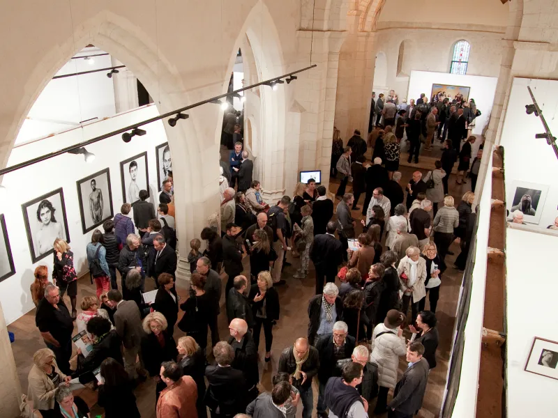 Crowd of visitors inside a historic stone church with vaulted ceilings, portrait exhibitions on walls, and stained glass windows.
