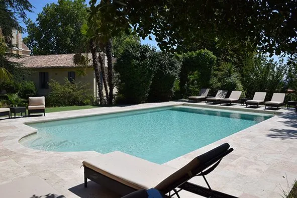 Rectangular swimming pool with turquoise water surrounded by lounge chairs and lush greenery, in front of a stone villa with paved terrace at Château Bellefont-Belcier in Bordeaux