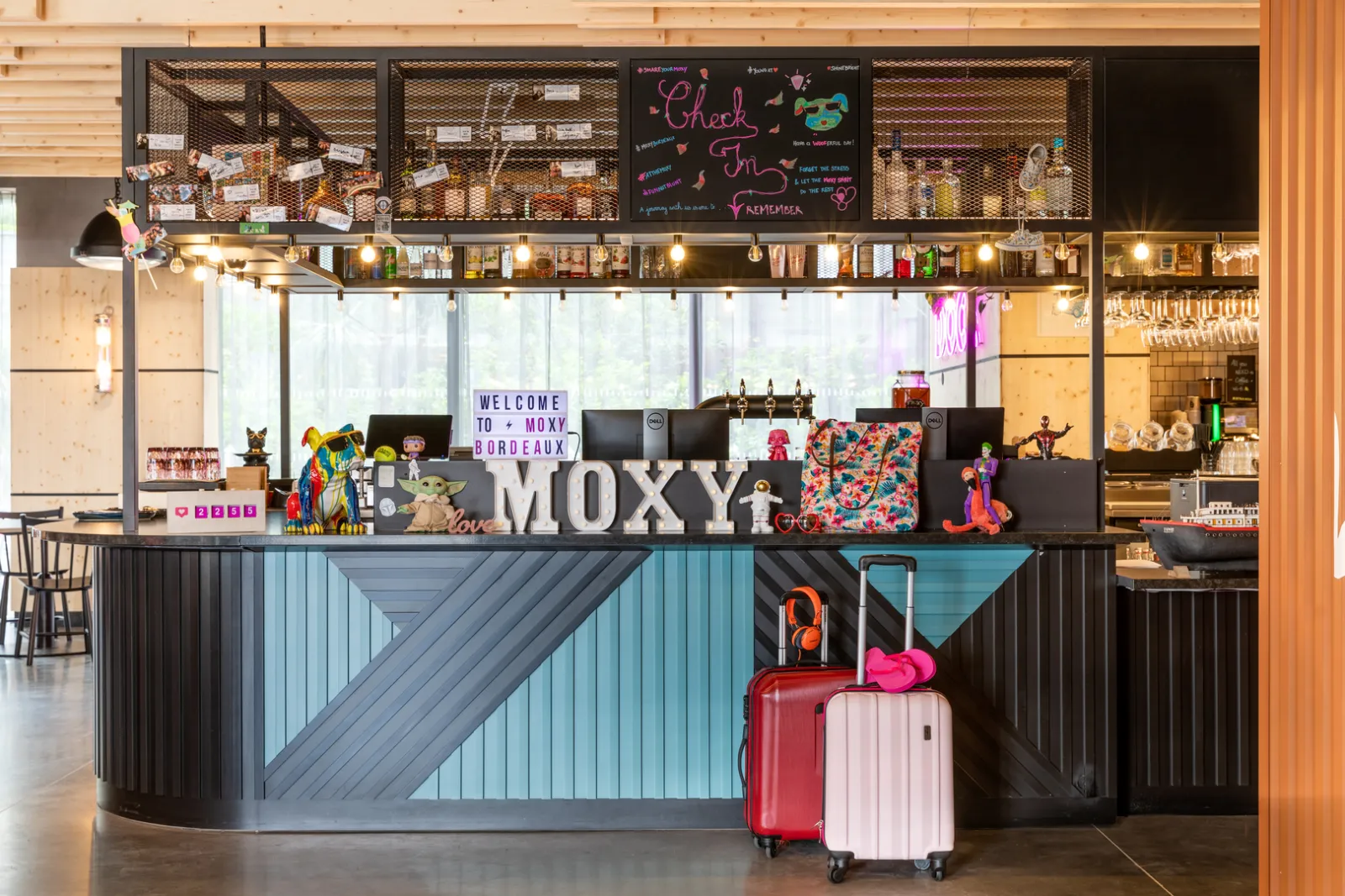 Moxy Bordeaux hotel reception desk with colorful luggage, welcome signage, and vibrant modern décor featuring turquoise and black accents