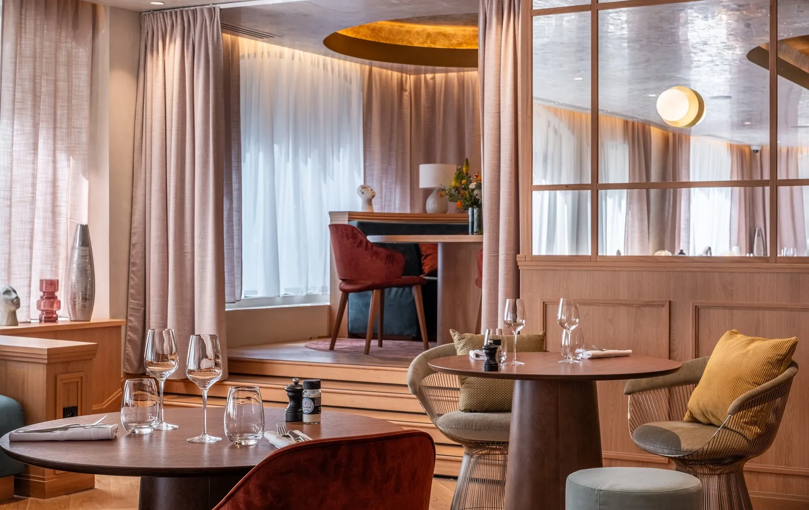 Madame B restaurant in Bordeaux featuring round wooden tables, modern chairs, large windows with beige curtains, warm golden and red decor, and lounge area in the background
