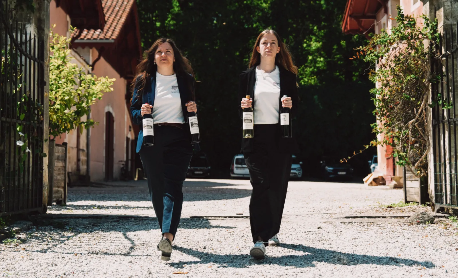 Two women walk down a cobblestone alley in Bordeaux, each holding two wine bottles from Château Picque Caillou, wearing black blazers and white t-shirts.