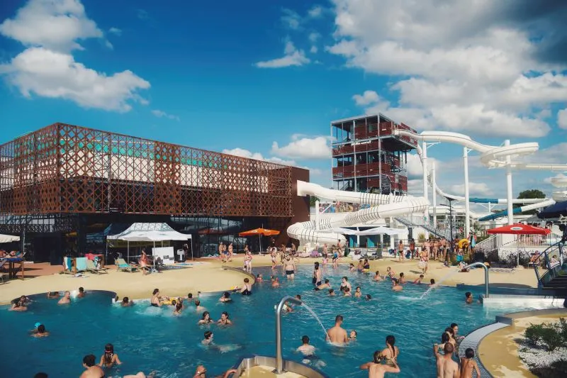 Aerial view of La Calinésie water park in Libourne featuring a large outdoor swimming pool with visitors, white water slides, modern metallic rust-colored building, sandy beach area, and clear blue sky.