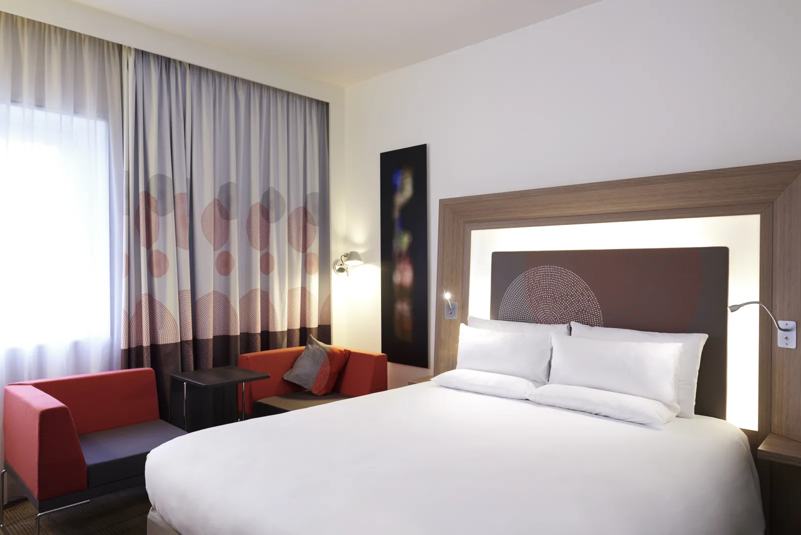 Modern hotel room at Novotel Bordeaux Lac featuring a king-size bed with white linens, a seating area with red armchair, gray and red curtains, and contemporary decor.