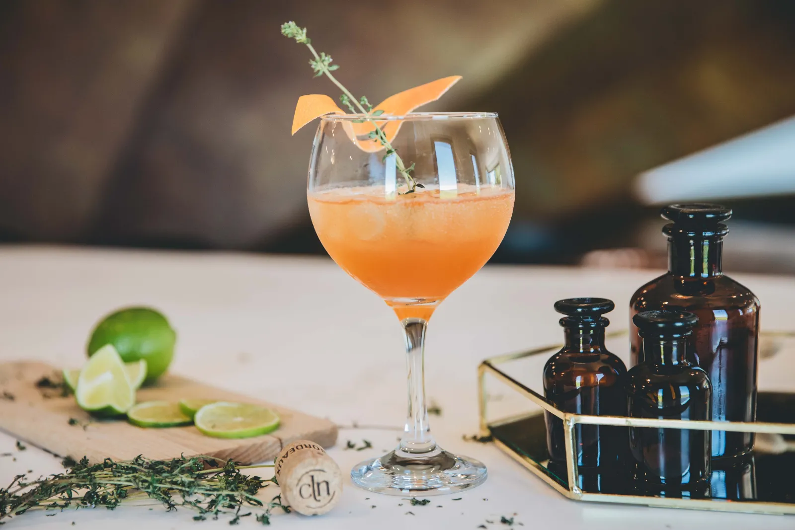 Orange cocktail in a glass with ice cubes, garnished with an orange slice and fresh thyme sprig, accompanied by sliced limes and dark brown spirit bottles on a white table.