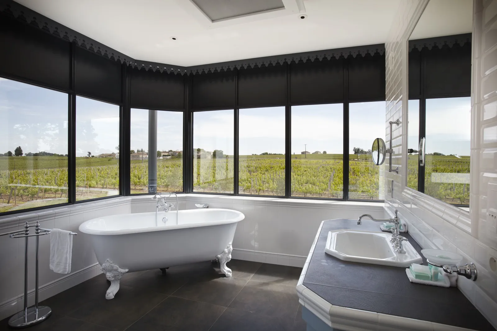 Luxury bathroom featuring a white freestanding bathtub, integrated sink, and panoramic windows overlooking Bordeaux vineyards