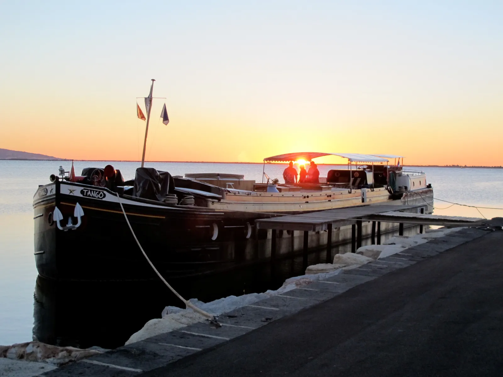The Tango boat moored at Bordeaux's quay at sunset, with its name visible on the black hull and flags flying from the bow.