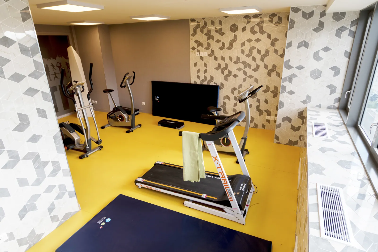 Modern fitness room with bright yellow flooring, cardio equipment including treadmill and stationary bikes, TV screen, geometric patterned walls in beige and white, at Mer & Golf City Bordeaux Lac Apartment Hotel