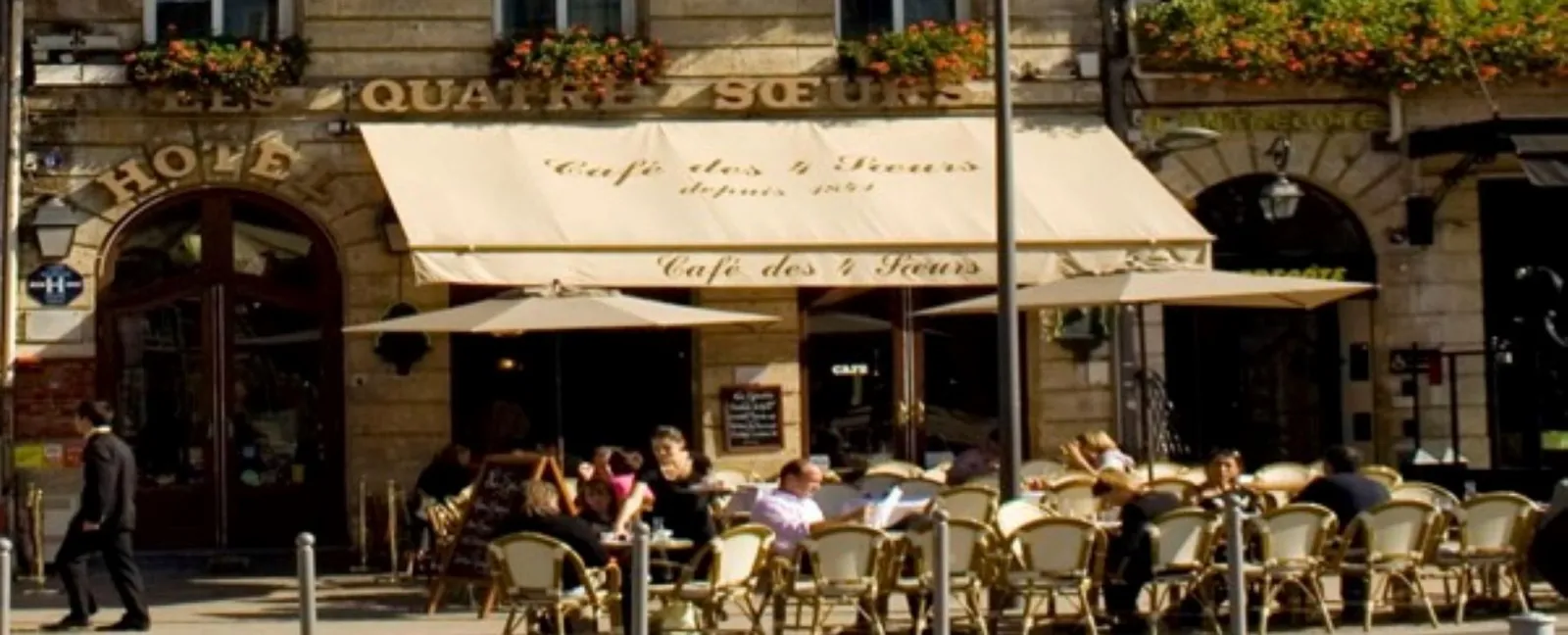 Sunny terrace of Café des 2 Fleurs in front of the Hôtel des Quatre Sœurs in Bordeaux, with customers seated at tables and historic stone façade.