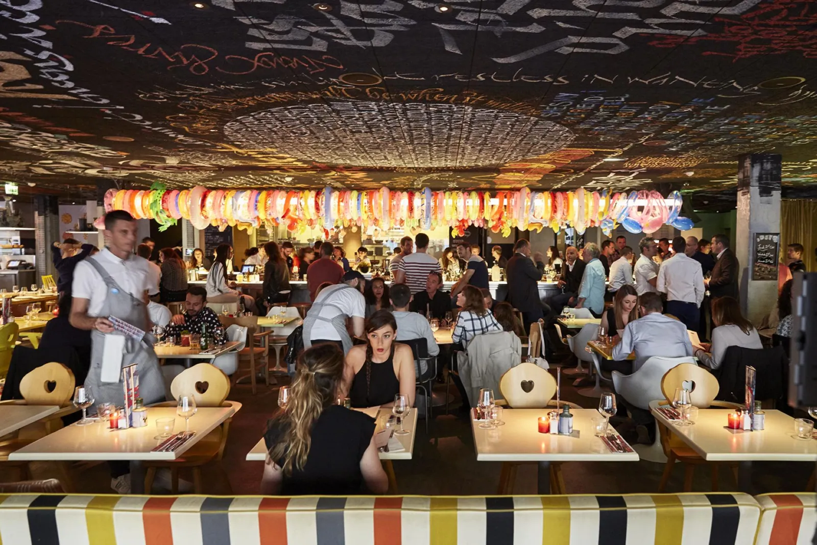 Interior view of Mama Shelter restaurant in Bordeaux with guests dining at tables, colorful ambient lighting and decorative lanterns hanging from the ceiling