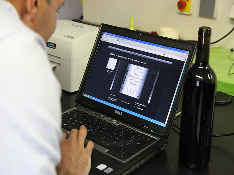 A man views a digitized wine bottle label on a Dell laptop screen, analyzing a Bordeaux wine in a tasting and evaluation context.