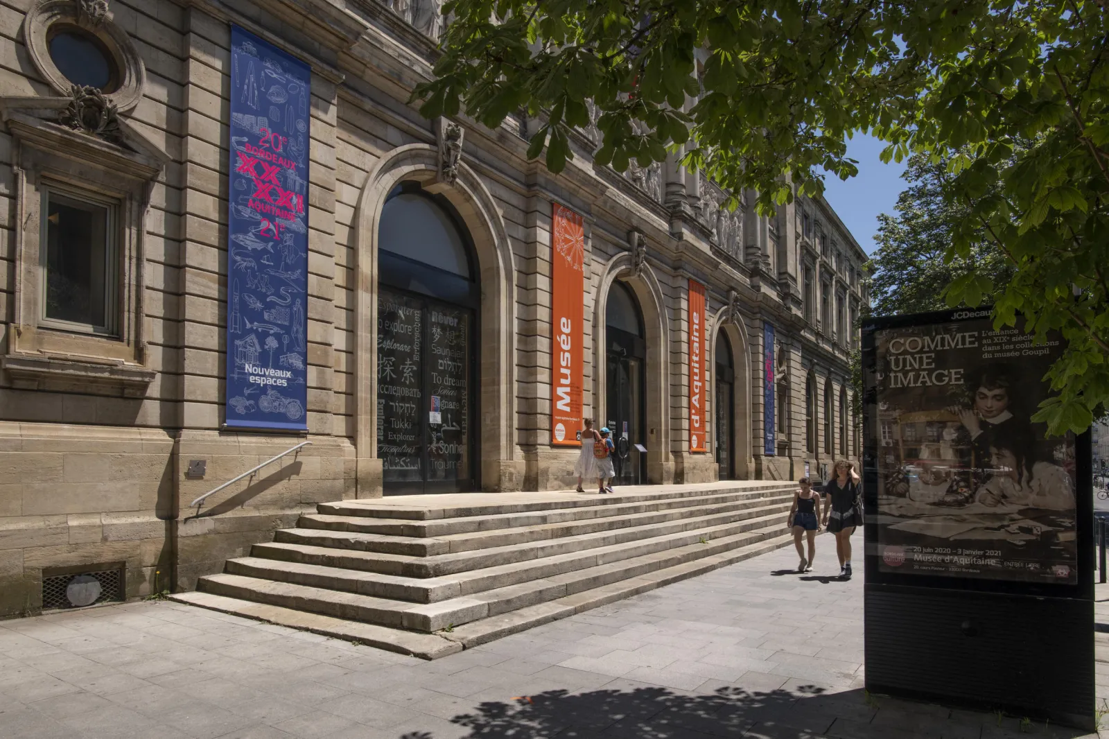 Facade of the Museum of Aquitaine in Bordeaux featuring large arched entrances, blue and orange exhibition banners, monumental steps, and visitors on a sunny day.