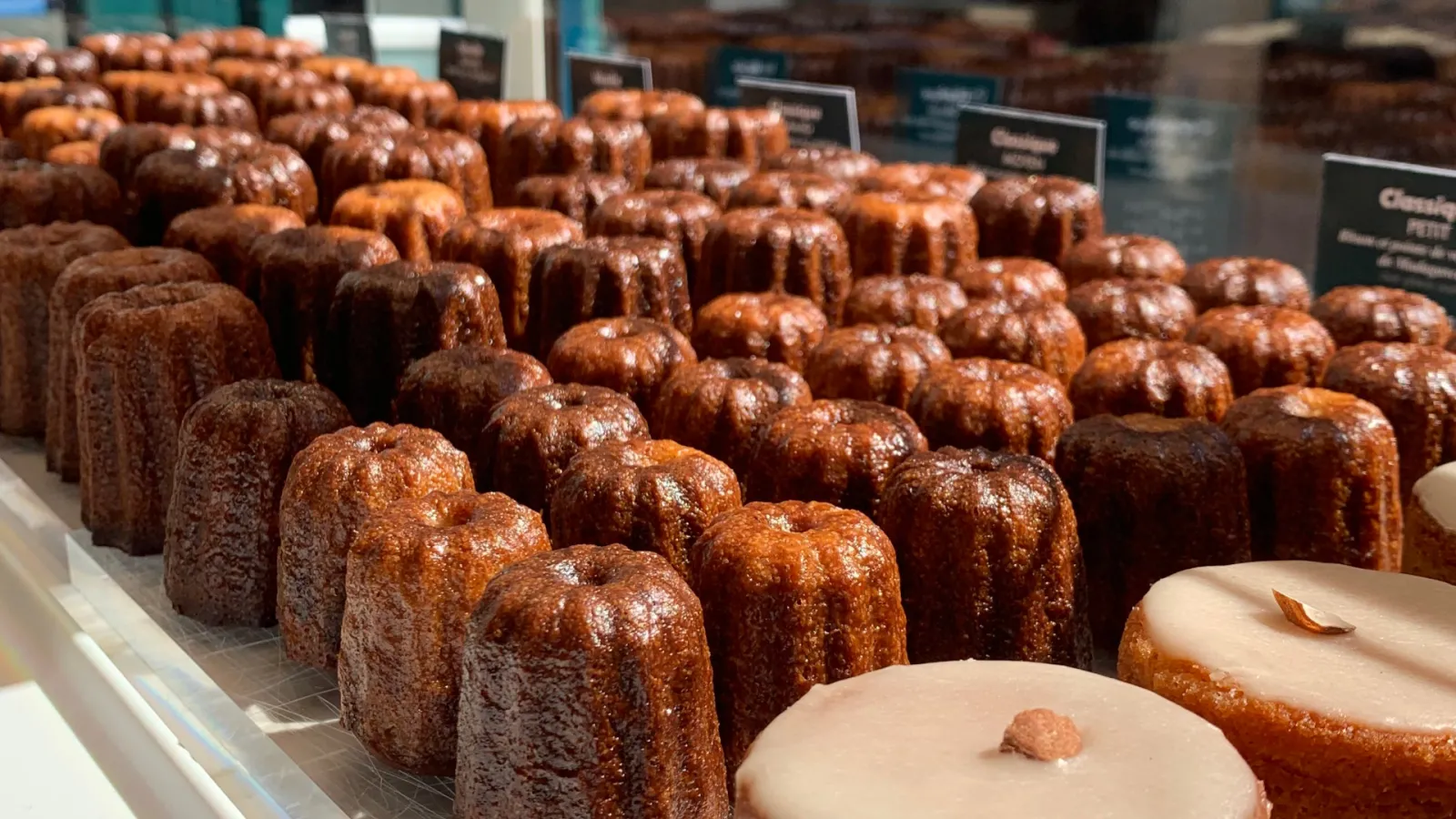 Pastry shop display case featuring golden caramelized cannelés and individually glazed cakes, traditional Bordeaux specialties