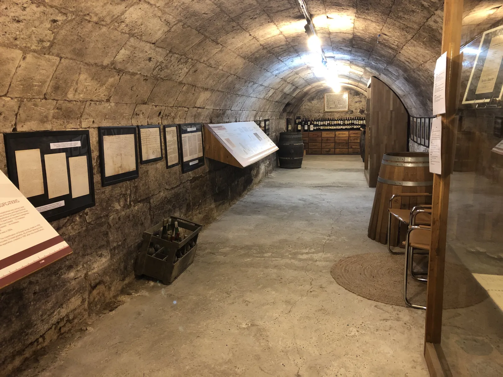Stone vaulted cellar interior with informational panels on the walls, wooden barrels, displayed wine bottles, and winemaking equipment at the Bordeaux Wine and Trade Museum.