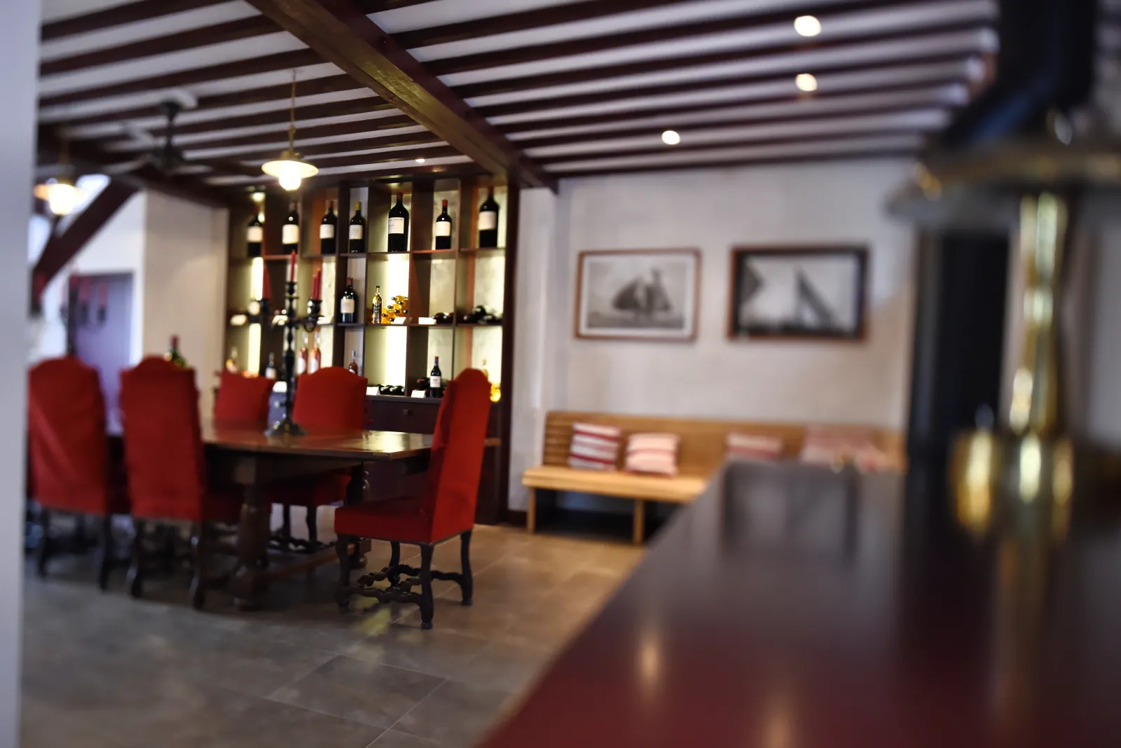 Elegant wine bar interior featuring a dark wooden counter with red chairs, illuminated wine bottle shelves, exposed beam ceiling and light wood furniture at Domaine de la Solitude in Bordeaux.