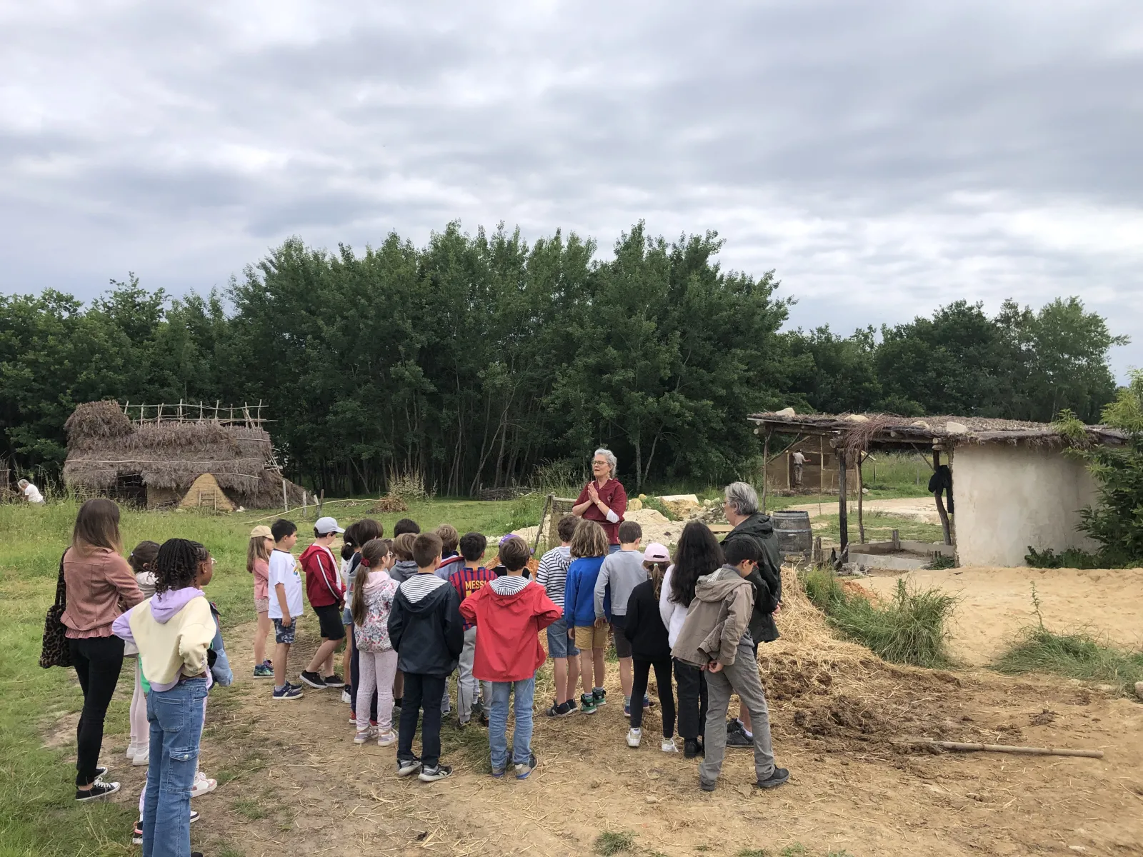 A group of children and adults listen to a guide during an educational tour at the medieval construction site of Guyenne in Bordeaux, with historical reconstructions and traditional buildings in the background.