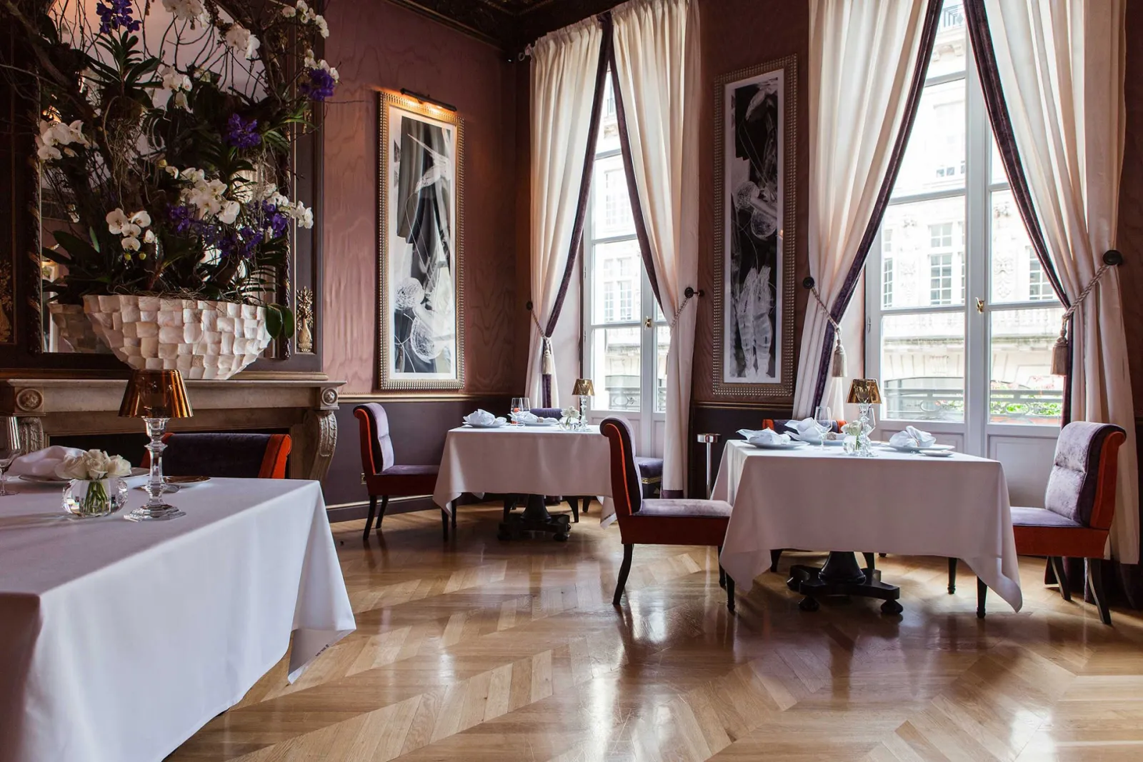 Elegant restaurant dining room featuring round tables with white tablecloths, burgundy chairs, tall windows with cream curtains, terracotta walls, ornate mirror and floral arrangements at Pressoir d'Argent Gordon Ramsay in Bordeaux.
