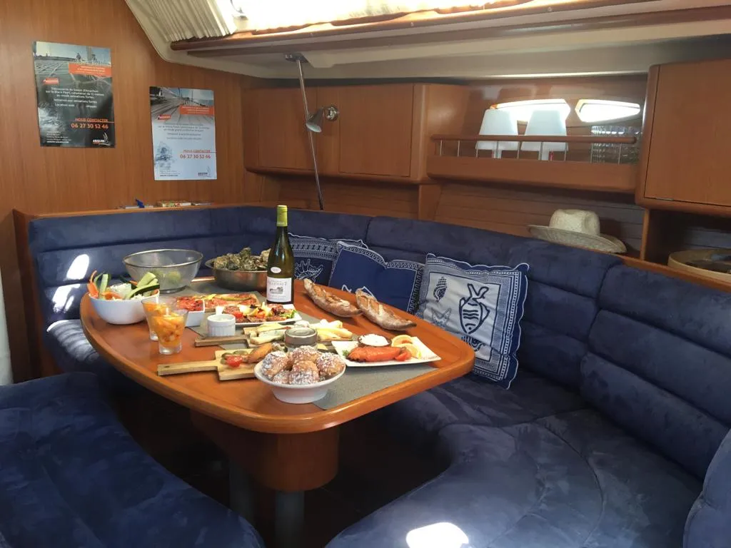 Interior of a sailing yacht featuring a dining table laden with gourmet food, wine, bread and cheese platter, surrounded by navy blue seating and teak woodwork.