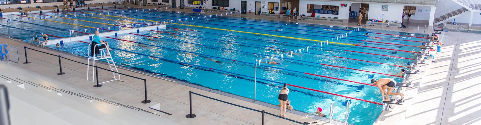 Aerial view of the UCPA Aqua Stadium in Mérignac, Bordeaux, featuring multiple training pools separated by colorful swim lanes, swimmers and divers in action, and modern facilities in the background.