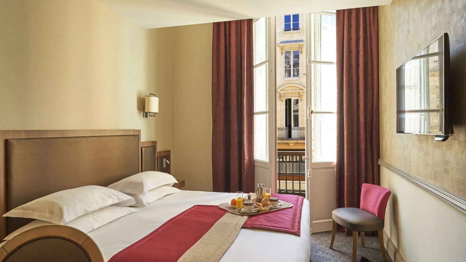 Elegant bedroom at Best Western Premier Hôtel Bayonne Etche Ona in Bordeaux, featuring a queen-size bed, light wood furnishings, burgundy curtains, balcony overlooking Haussmann architecture, wall-mounted TV and pink chair.