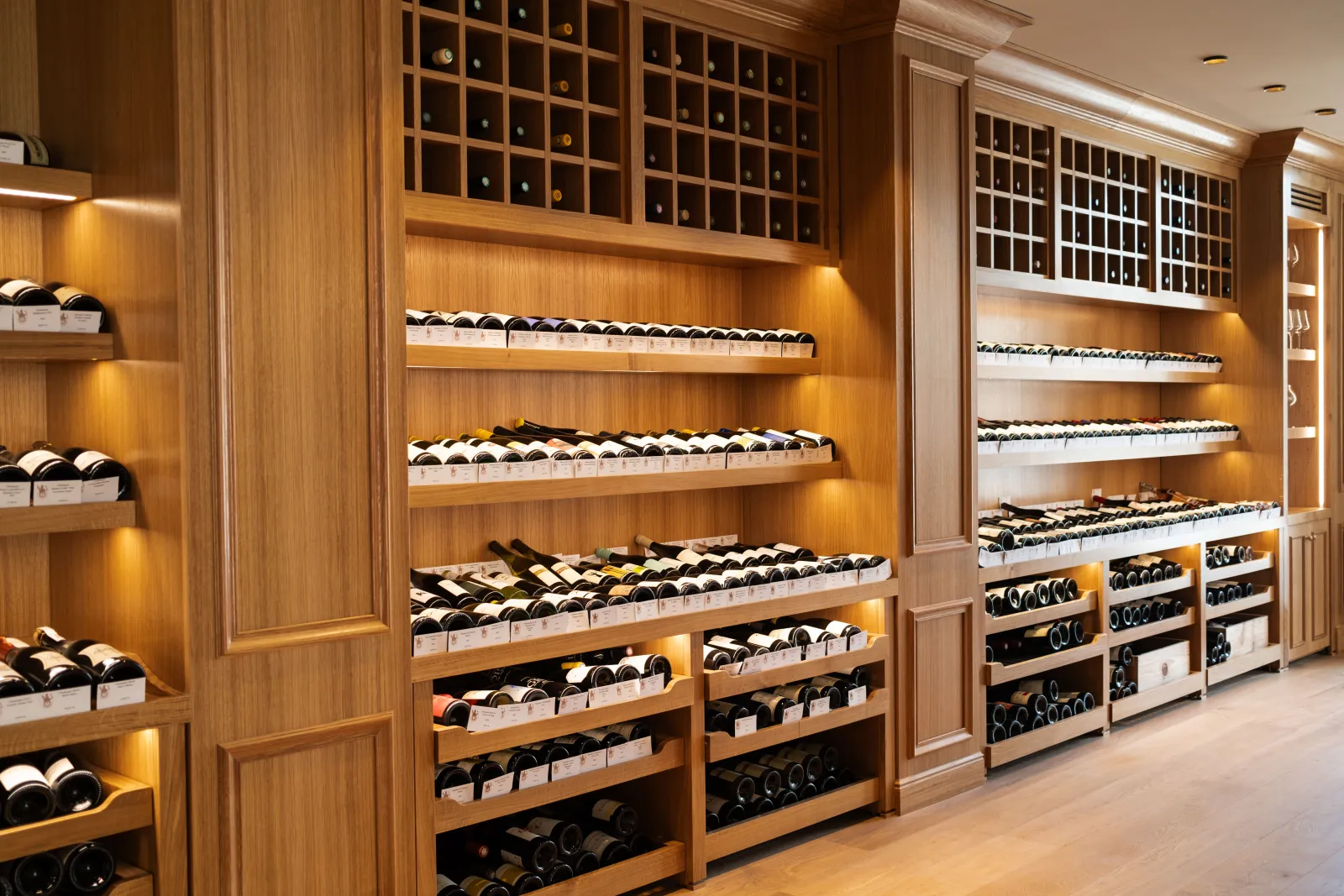 Elegant wine cellar featuring bottles displayed on light wood shelves and storage racks with warm golden lighting, showcasing a sophisticated Bordeaux wine collection.