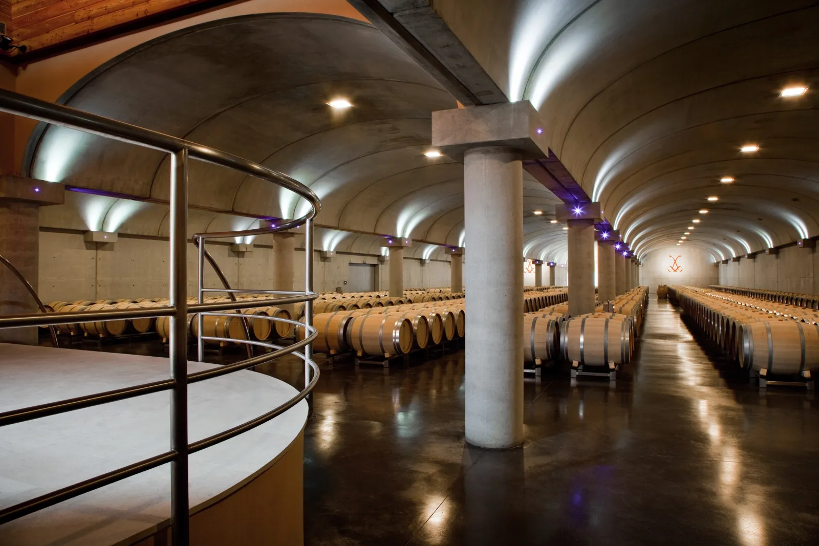Vaulted wine cellar of Château La Louvière in Bordeaux featuring rows of wooden barrels beneath an arched concrete ceiling with recessed lighting and support columns.