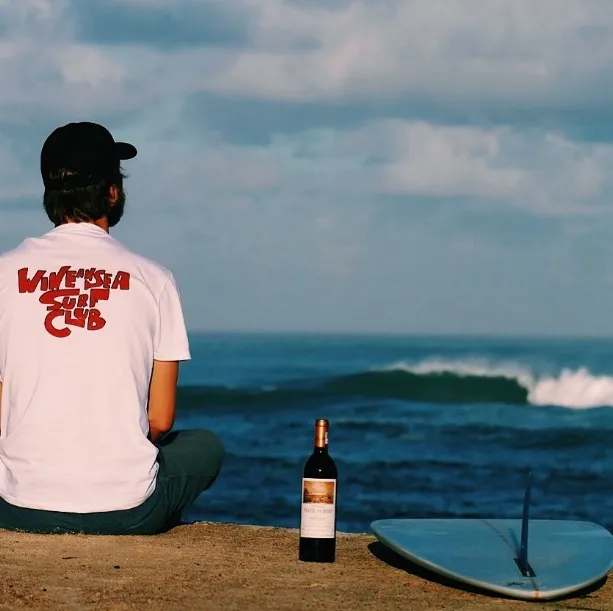 A man sitting on the sand facing the ocean, wearing a white t-shirt with red logo, a bottle of Bordeaux wine placed beside him, and a blue surfboard visible on the beach.