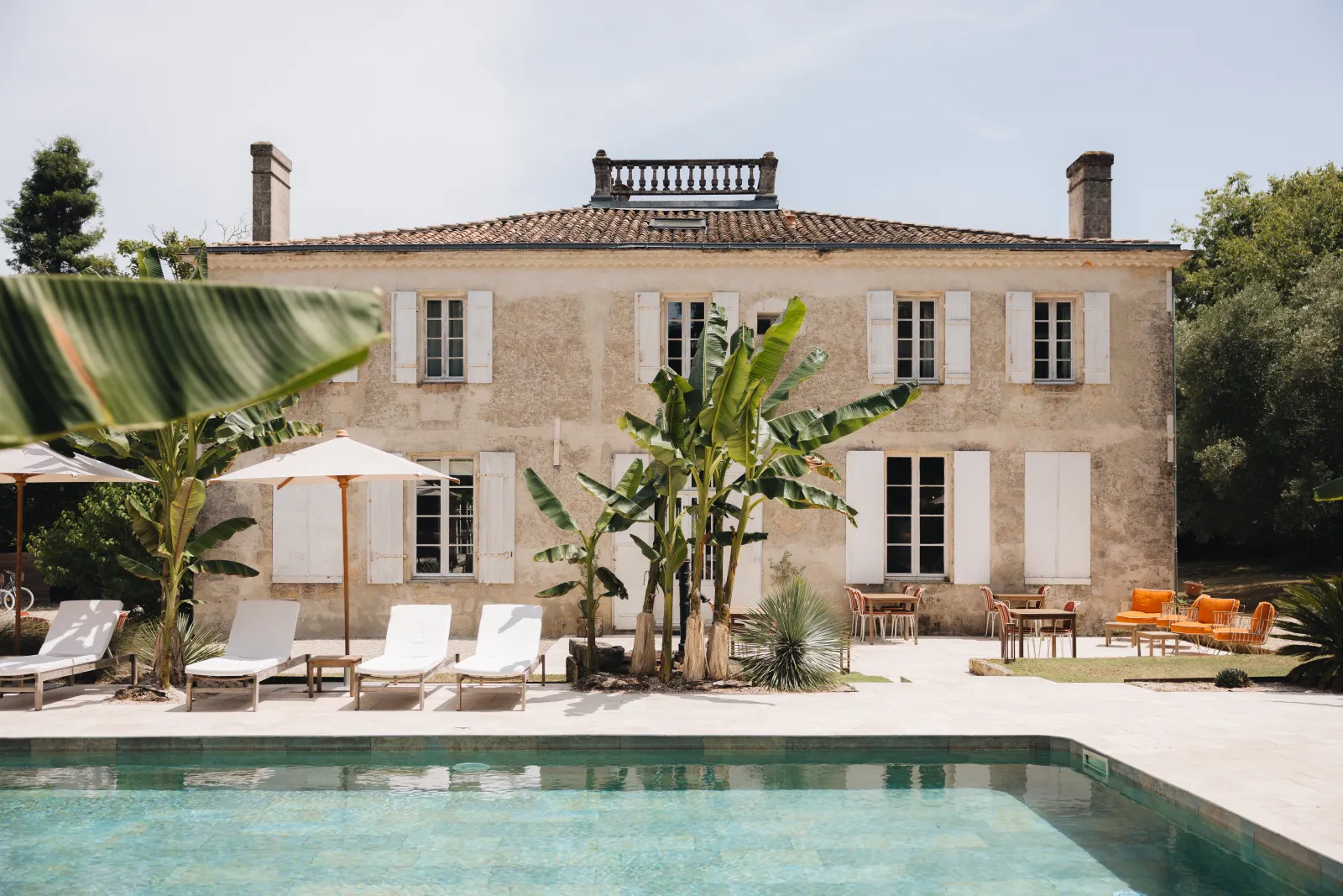 Front view of Château Réal in Bordeaux featuring a stone facade with white shutters, a turquoise swimming pool in the foreground, sun loungers and umbrellas, surrounded by Mediterranean plants.