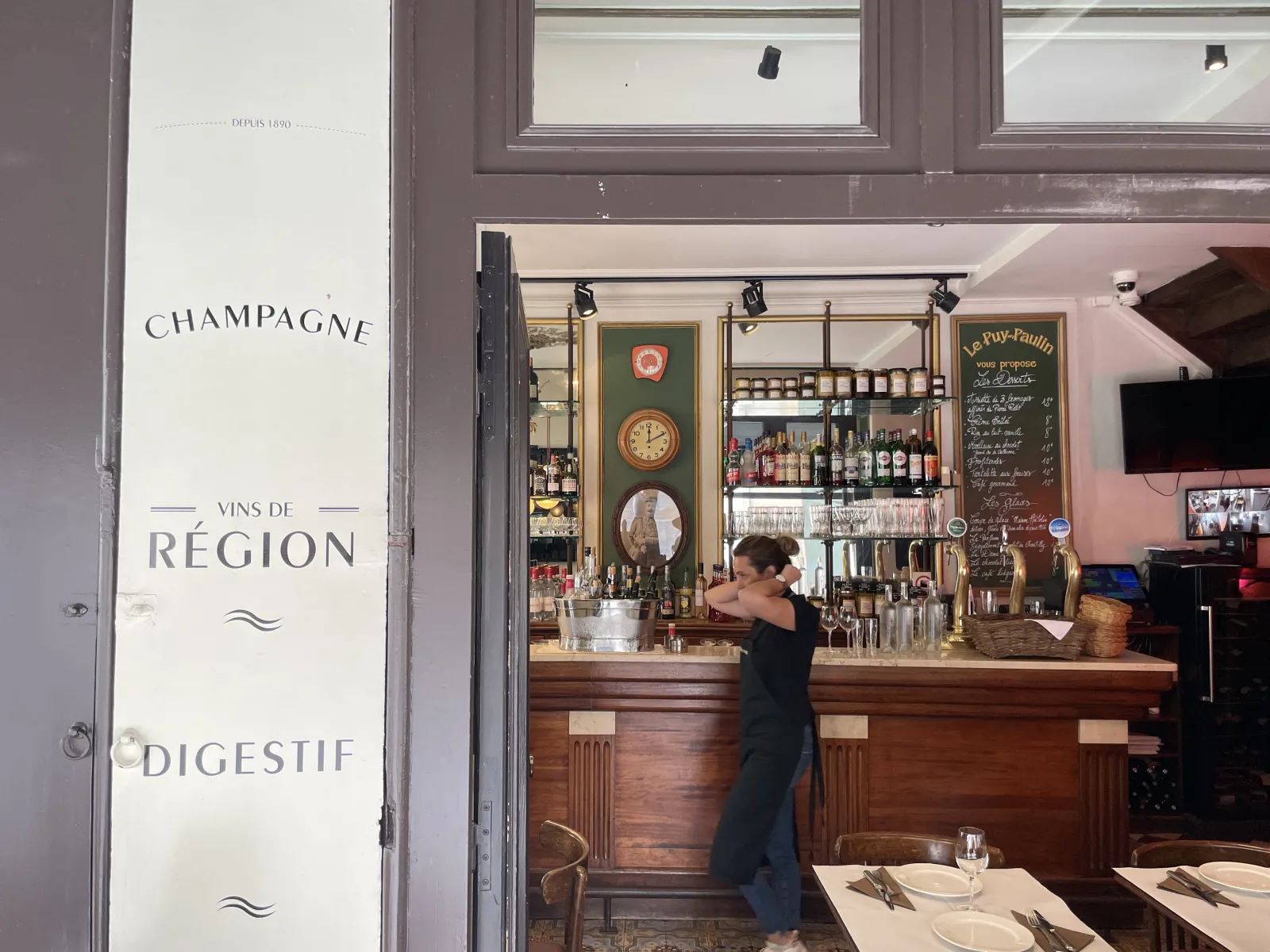 Interior view of Le Puy-Paulin restaurant in Bordeaux, featuring a bartender at the wooden counter, displayed bottles of champagne and regional wines, and an entrance door showcasing house specialties.
