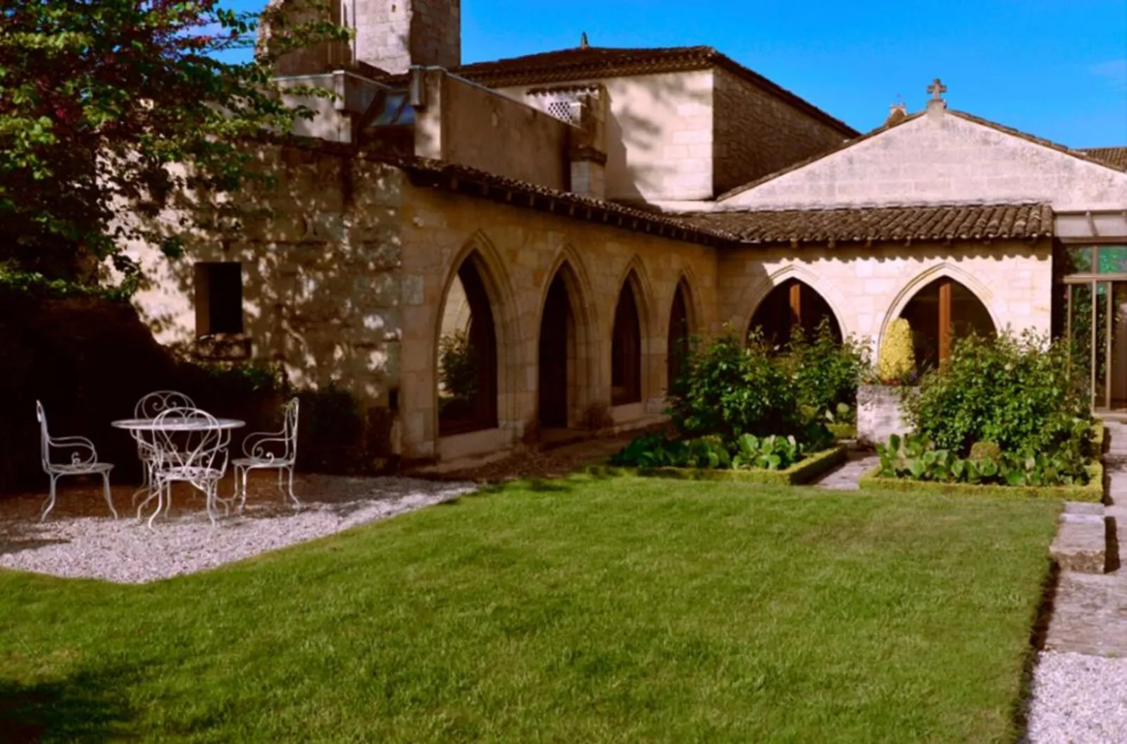 Interior courtyard of the Jacobins Convent in Bordeaux featuring Gothic architecture, pointed arches, green lawn, wrought iron garden furniture, and historic buildings under clear blue sky.