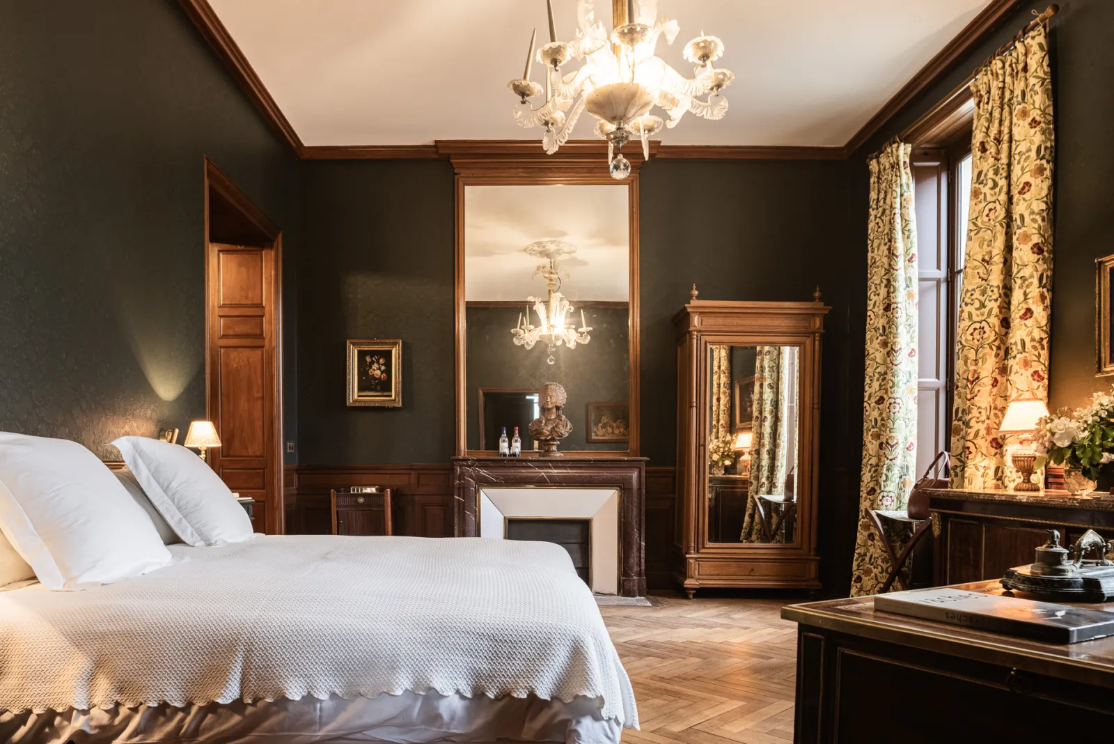 Luxurious guestroom at Château Sénéjac in Bordeaux, featuring a white bed with pillows, charcoal gray walls with wood paneling, crystal chandelier, framed mirror, fine wood furniture, gold floral curtains, and classic elegant décor.