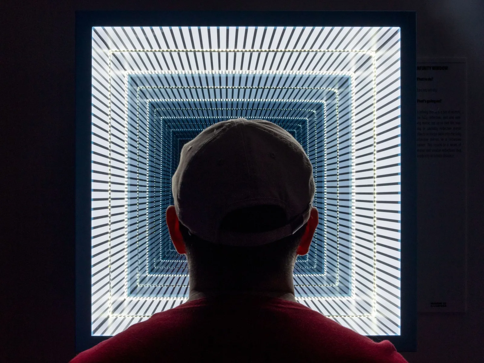 A person from behind wearing a red shirt facing an illuminated installation with white lines creating an infinite tunnel perspective effect at the Museum of Illusion in Bordeaux.
