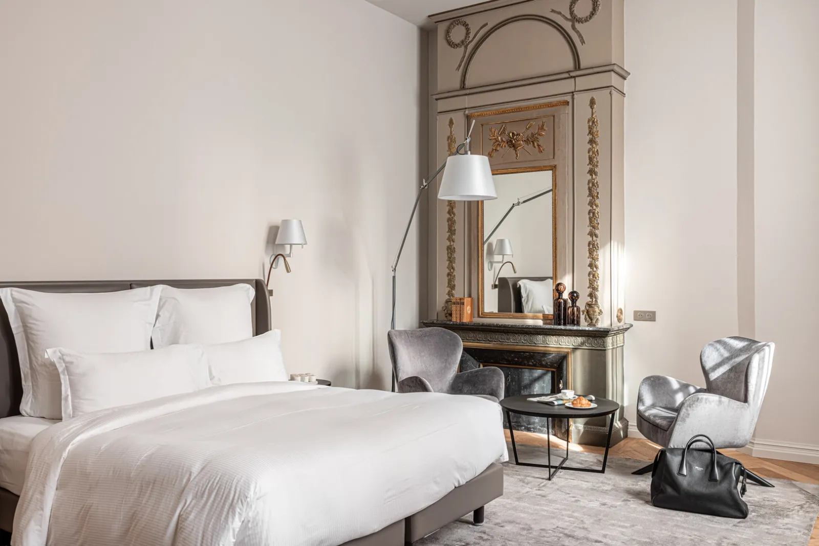 Elegant bedroom at Villas Foch hotel in Bordeaux featuring a king-size bed with white linens, ornate mirror above fireplace, gray armchairs, modern arc lamp, and refined classic décor