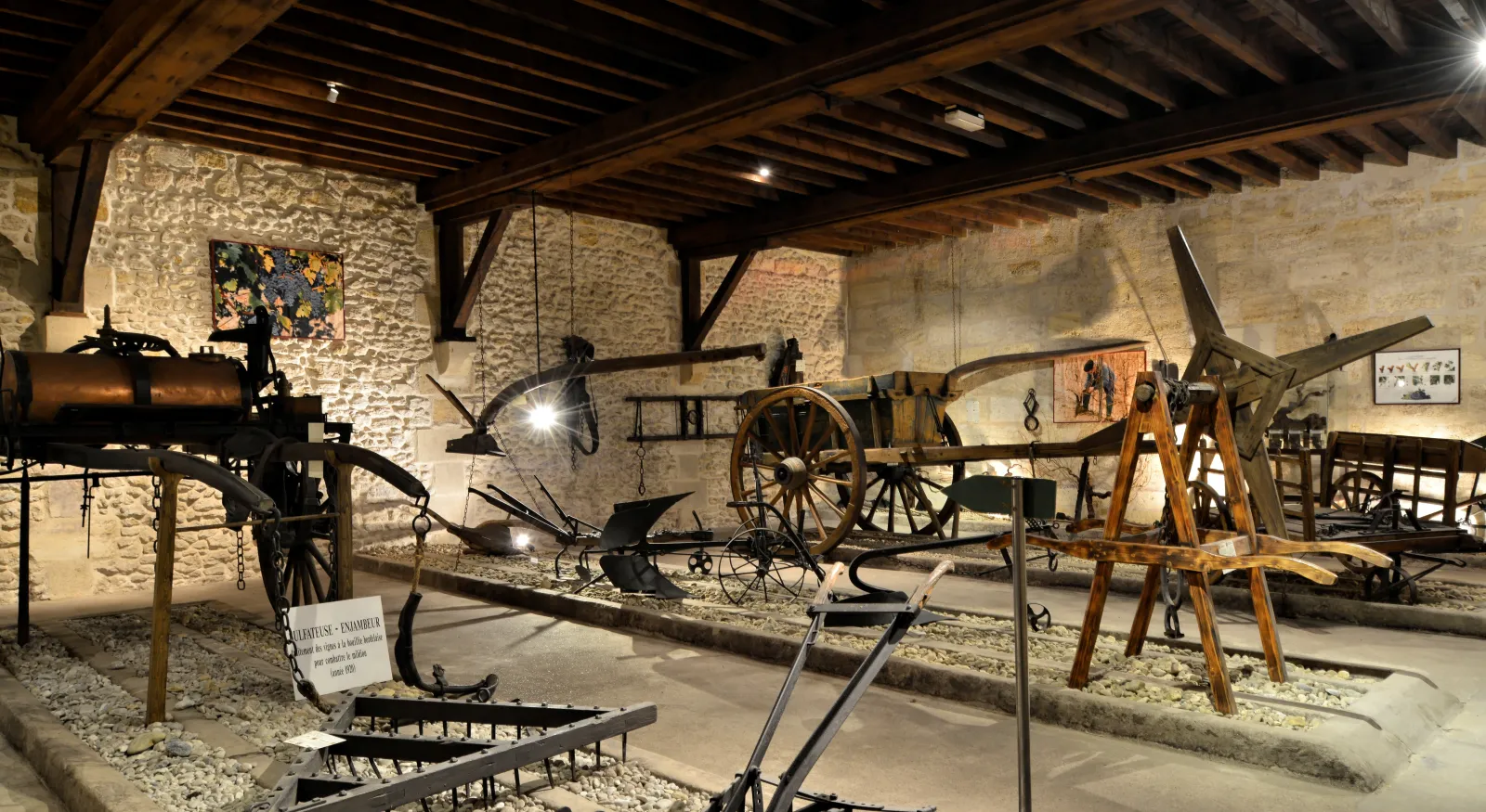 Interior of the Museum of Arts and Crafts of Wine and Vine in Bordeaux, displaying a collection of antique agricultural tools and machinery for viticulture, including plows, barrels, and wooden and metal equipment, exhibited under wooden beams with stone 
