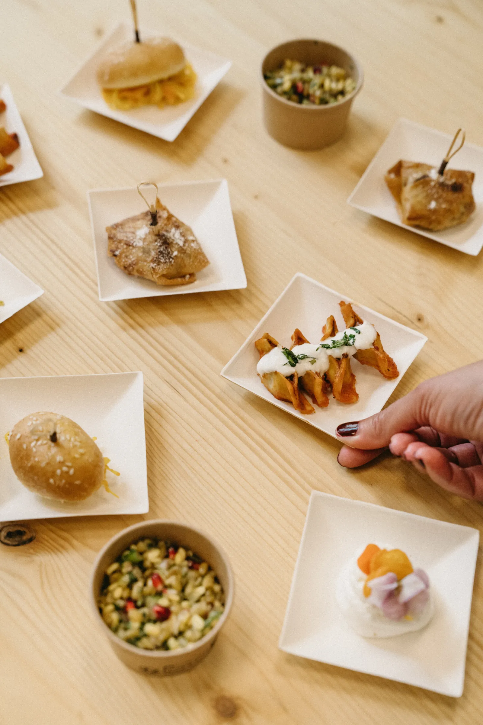 Selection of small plates and tapas from Marie Curry restaurant in Bordeaux, presented on white plates and bowls, featuring fritters, topped fries, mixed salad, and desserts, arranged on a light wooden table with a hand reaching for a plate.