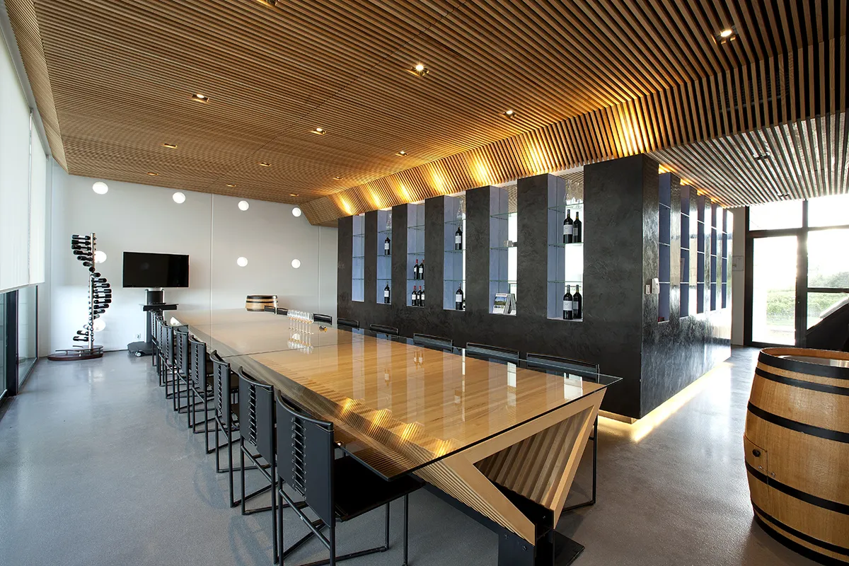 Modern tasting room at Château Faugères featuring a wooden counter with bar stools, wine bottle display cases, slatted wood ceiling with warm lighting, and traditional oak barrels.