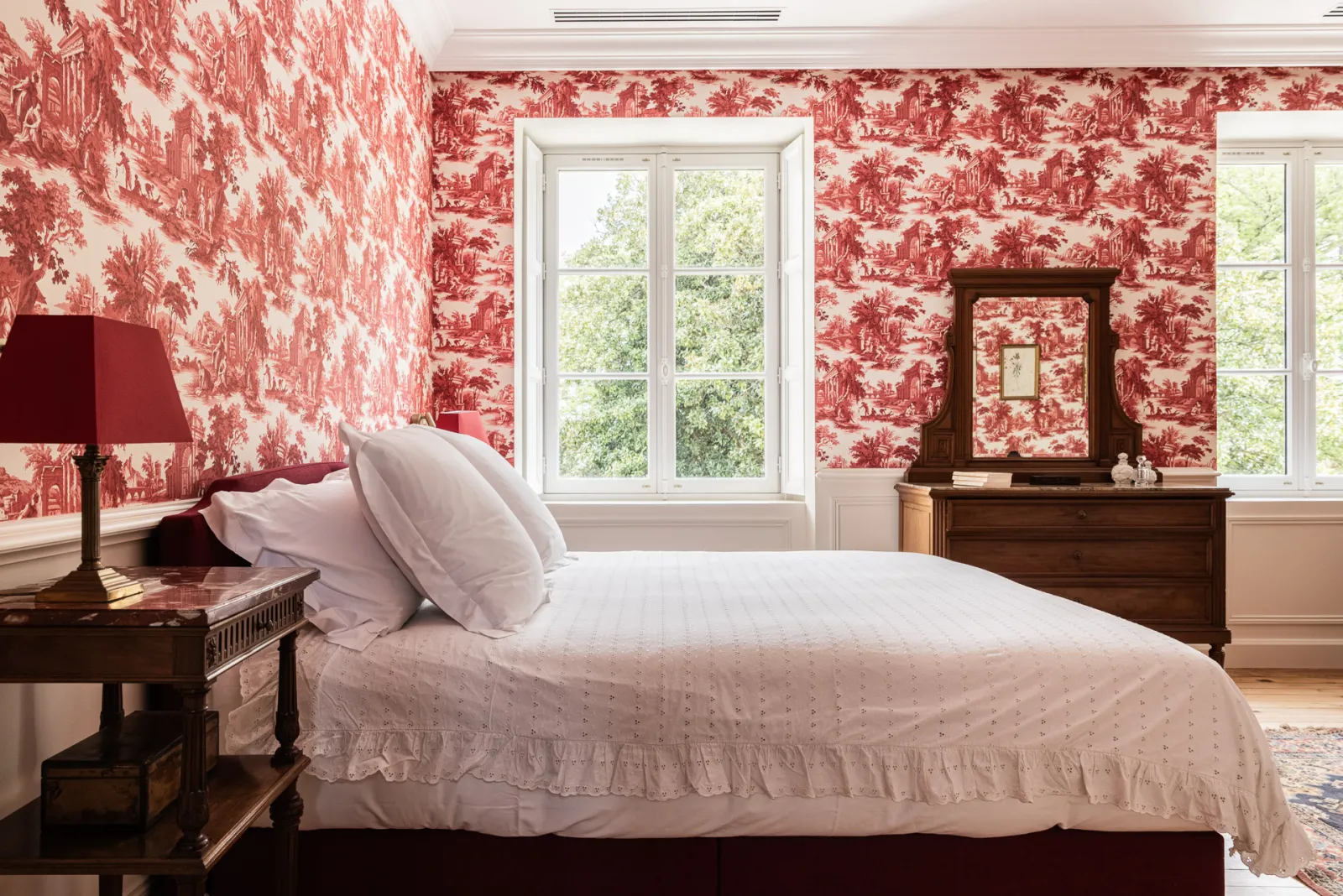 Elegant guest bedroom featuring red toile de Jouy wallpaper, wooden bed with white linens, antique furniture, bedside lamp, gilt mirror and large windows overlooking greenery.