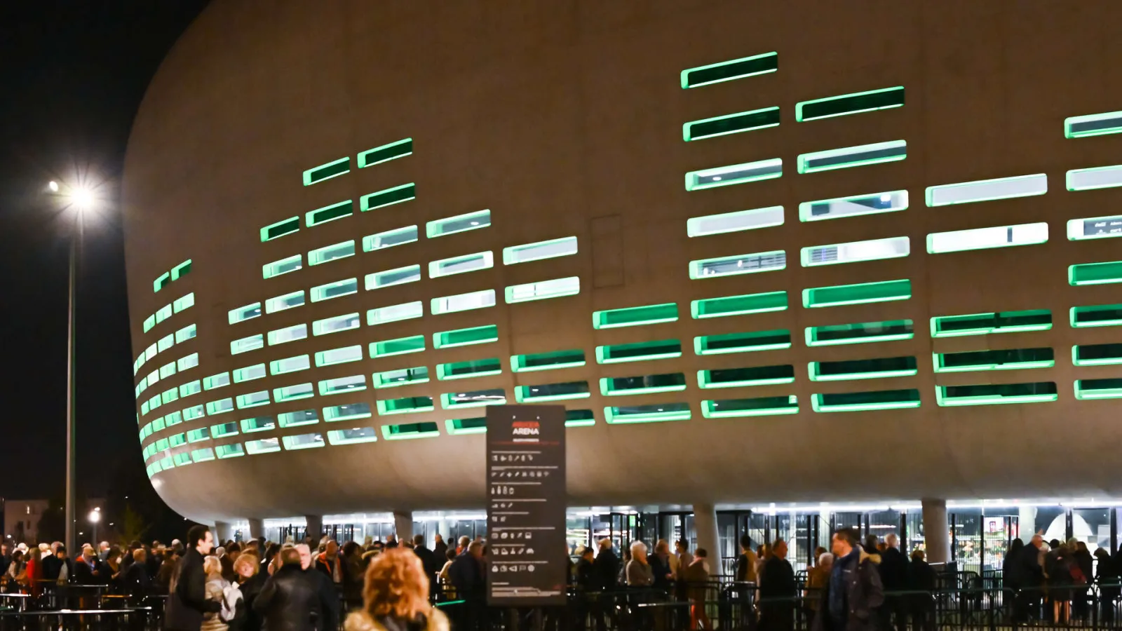 Night view of Arkea Arena in Floirac, Bordeaux, featuring a green and white neon-lit curved facade with crowds of visitors below