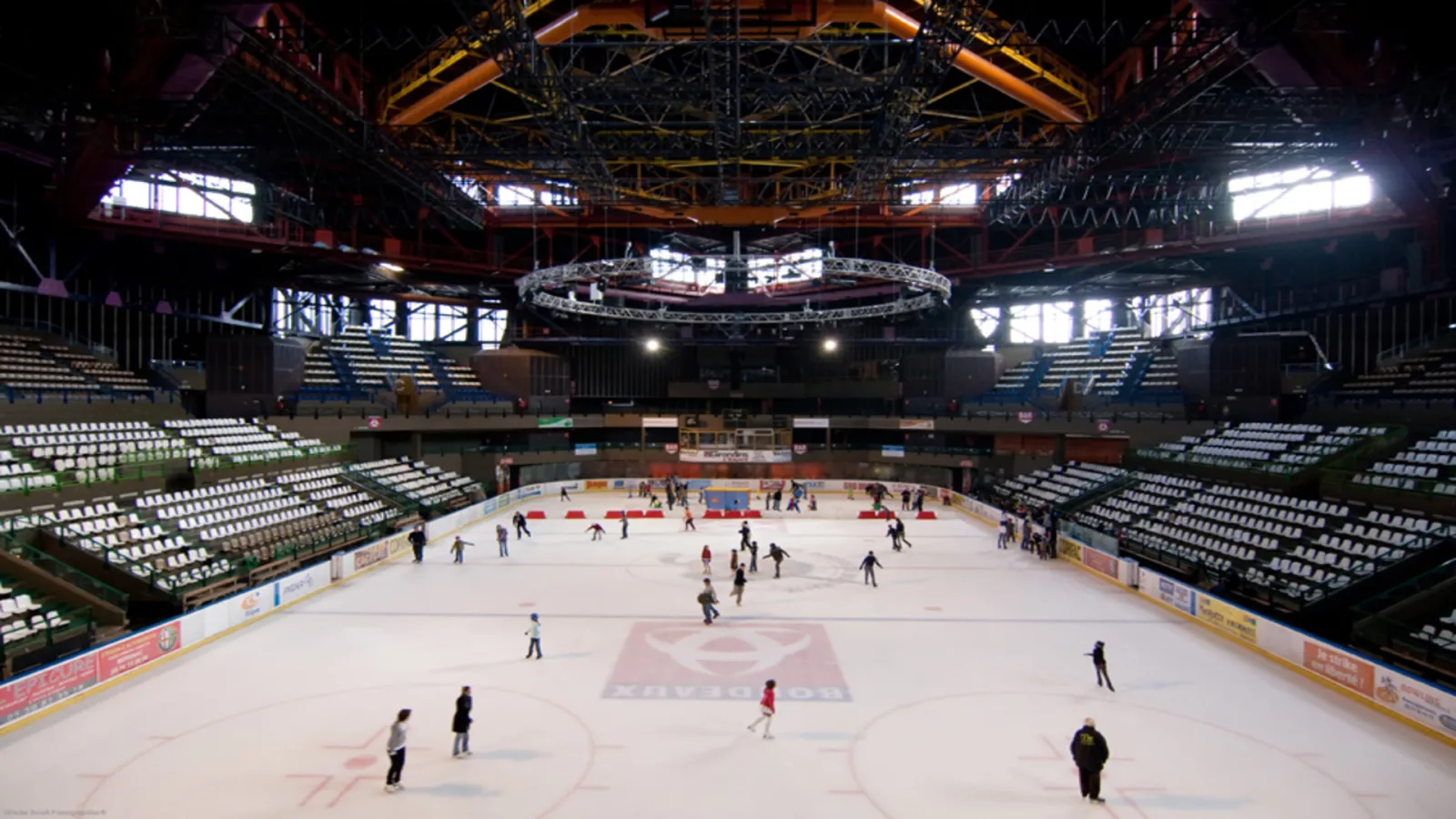 Interior view of Mériadeck Ice Rink in Bordeaux with multiple skaters on the ice, empty stands, and metal roof structure with orange and yellow lighting.