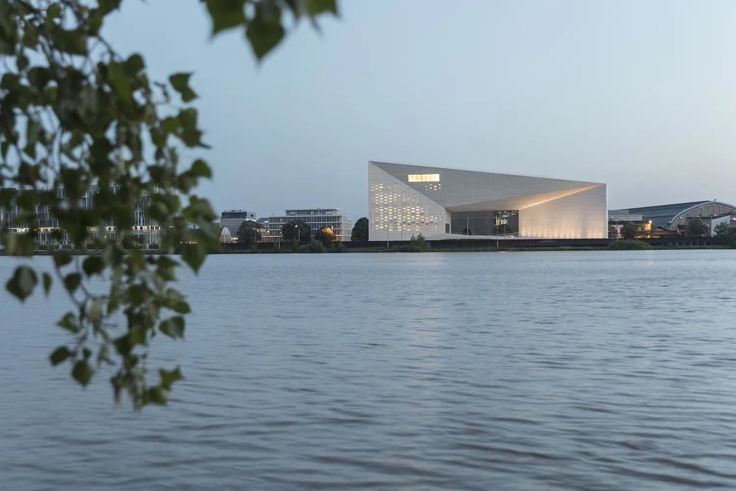 The FRAC Nouvelle-Aquitaine MECA building in Bordeaux, a modern architectural structure with clean geometric shapes, lit up at dusk, located on the banks of the Garonne River with the city in the background.