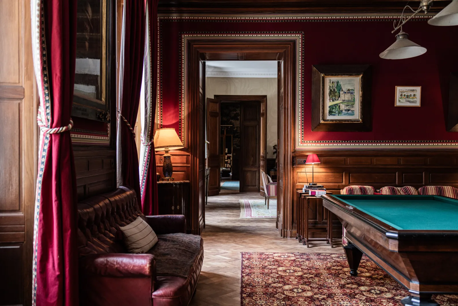 Elegant lounge room at Château Sénéjac featuring burgundy walls, Chesterfield leather furniture, billiard table with turquoise felt, solid wood doors, red curtains and warm lighting