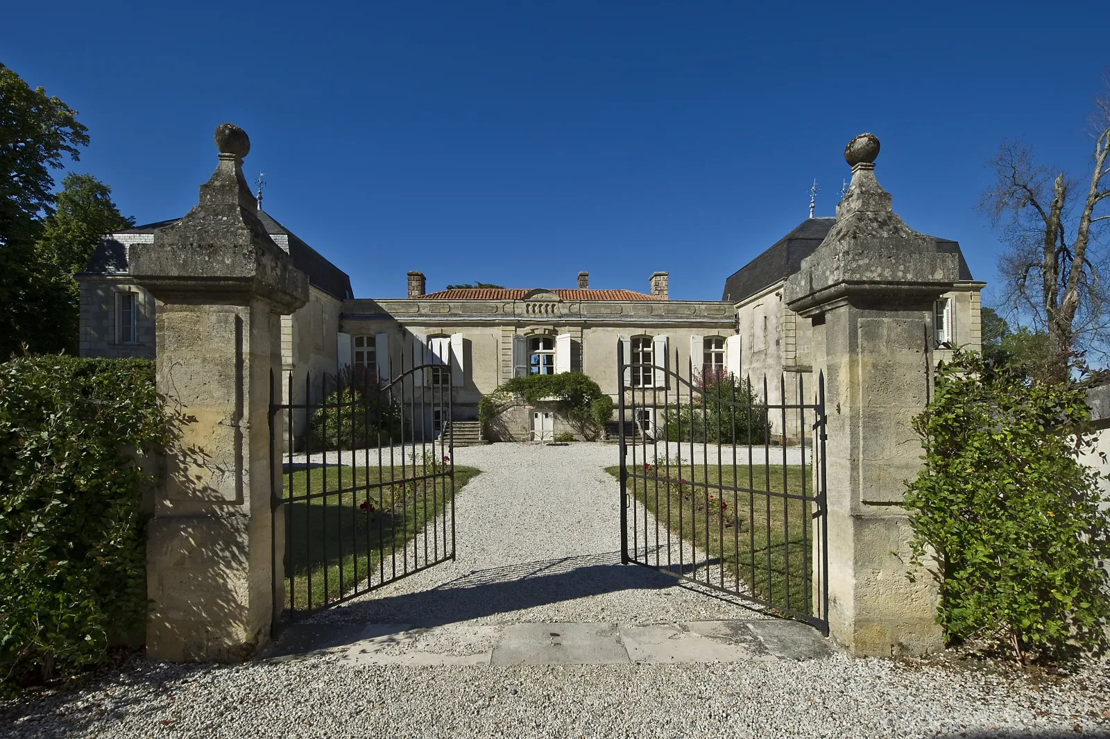 Front view of Château Picque Caillou in Bordeaux, a classical stone mansion with an ornate wrought-iron double gate, flanked by two symmetrical pavilions with mansard roofs, set against a clear blue sky.