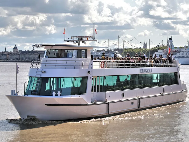 The Burdigala II cruise ship sailing on the Garonne River in Bordeaux, with passengers on the upper deck and the historic city skyline in the background.