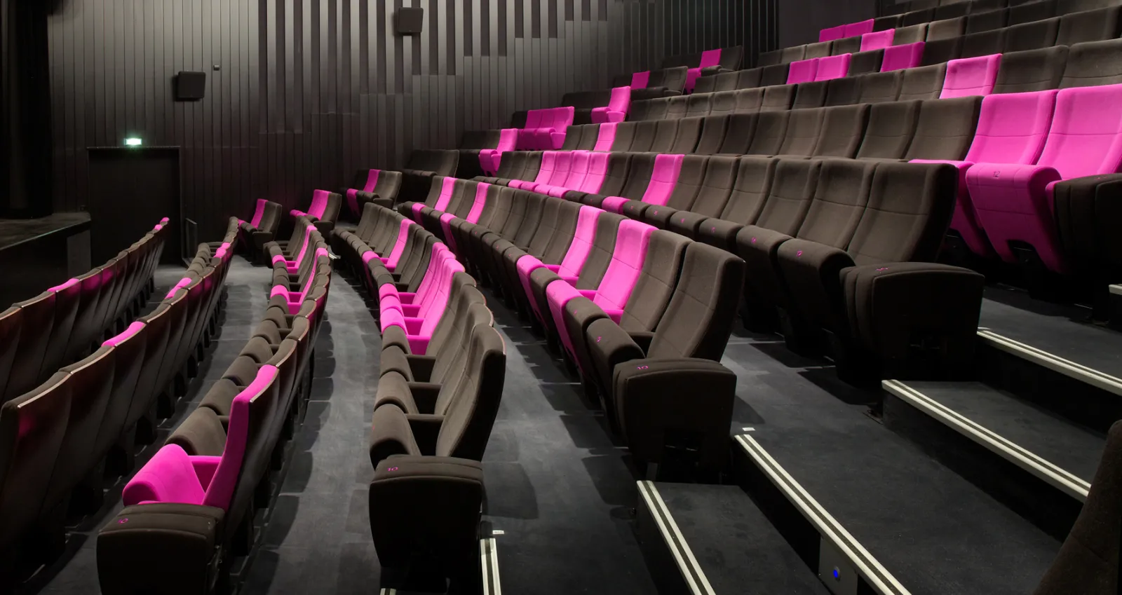 Modern cinema interior with black and hot pink recliners arranged in rows, pink architectural lighting on armrests, grey striped walls, and visible stairs in the background at Blanquefort, Bordeaux.