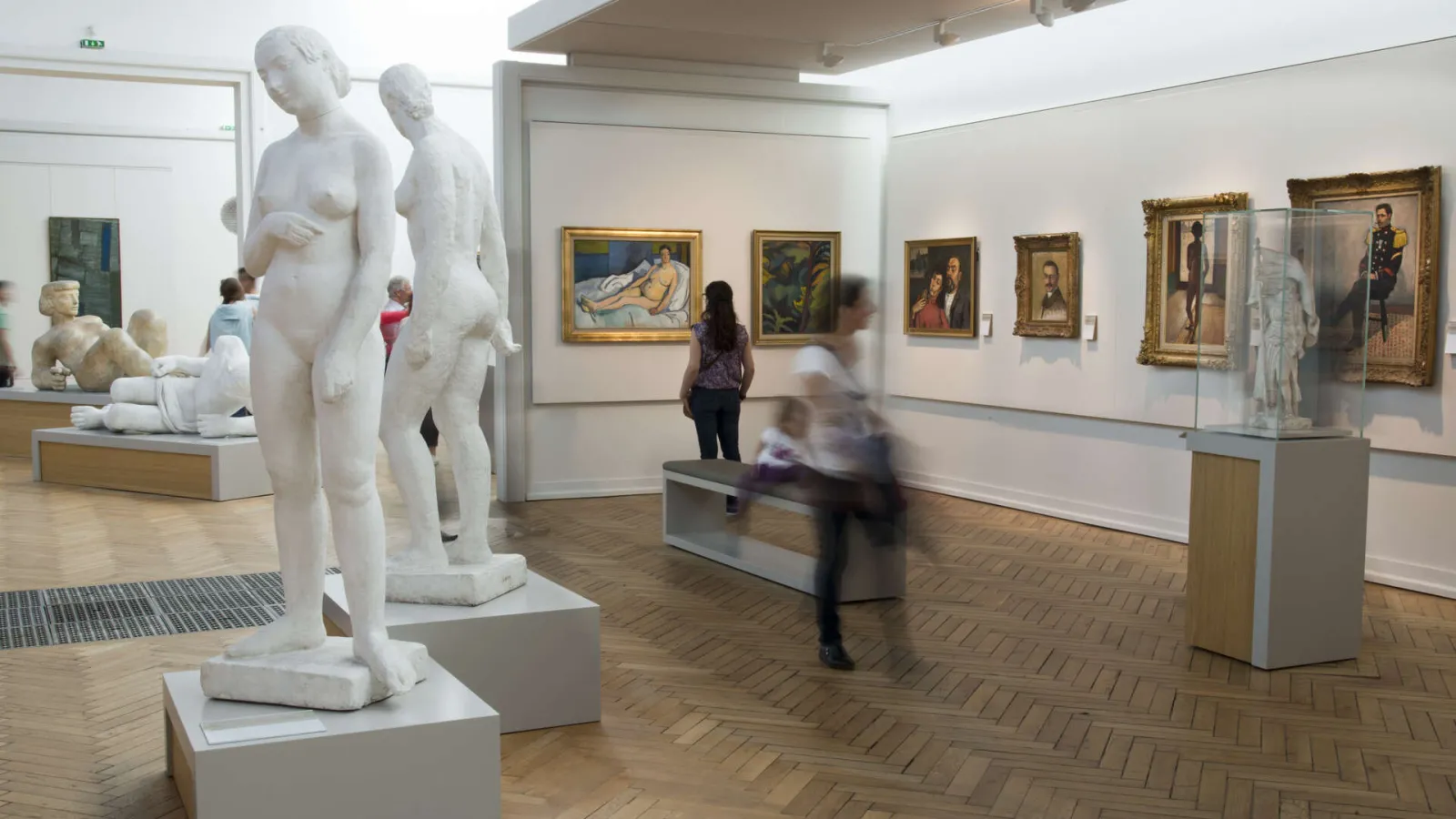 Gallery of the Bordeaux Museum of Fine Arts displaying classical white marble sculptures in the foreground and framed portrait paintings on the walls, with visitors moving through the exhibition space.