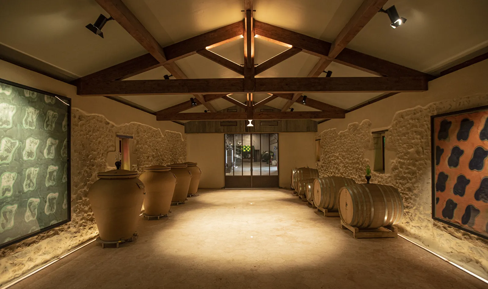 Interior of a winemaking cellar at Domaine des Perchés in Bordeaux, featuring fermentation vats, aging barrels, and display cases under exposed wooden beams.