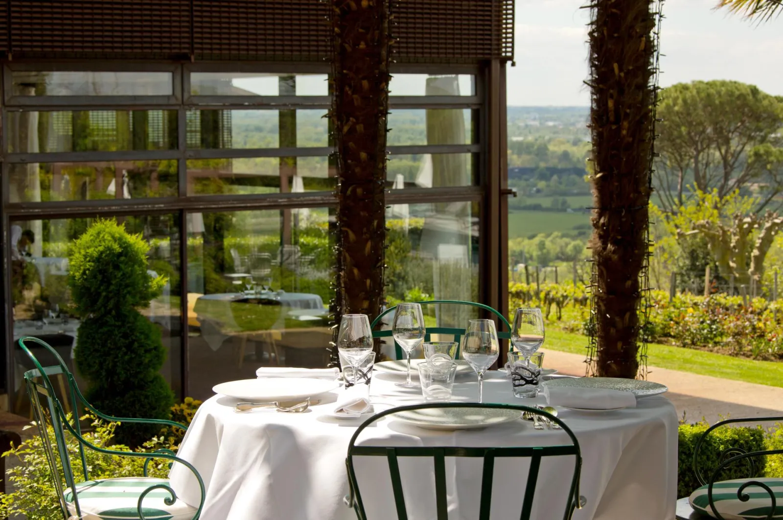 Fine dining table set with white tablecloth, glassware, and place settings on a terrace overlooking Bordeaux vineyards and valley landscape, framed by stone columns and large windows.
