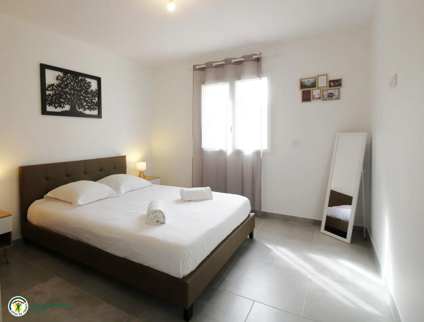 Bright and modern bedroom in the Tanins guesthouse at Château Pierre de Montignac in Bordeaux, featuring a double bed with white linens, gray headboard, window with natural light, standing mirror, and minimalist decor.