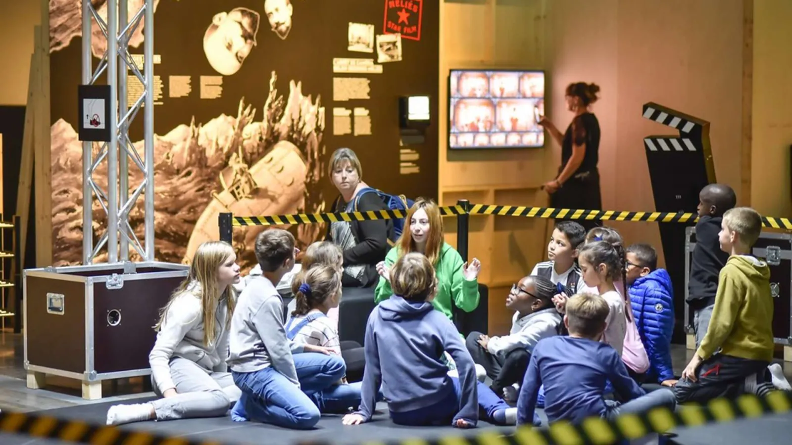 A group of children sitting on the floor participate in an interactive activity at a science museum, with educators and exhibitions about cinema and technology in the background.